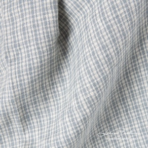 May include: Close-up of a light blue and white checkered fabric. The fabric has a textured appearance with a subtle pattern. The image shows the fabric draped, with soft folds and shadows. The fabric is likely linen.