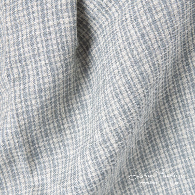 May include: Close-up of a light blue and white checkered fabric. The fabric has a textured appearance with a subtle pattern. The image shows the fabric draped, with soft folds and shadows. The fabric is likely linen.