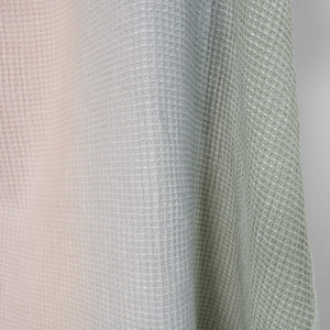 Può includere: Primo piano di un tessuto strutturato con un motivo a nido d'ape. Il tessuto passa da una tonalità rosa pallido a sinistra a una tonalità verde chiaro a destra, con un grigio neutro al centro. Il materiale appare morbido e flessibile.