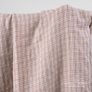 May include: A close-up shot of a folded fabric featuring a small, checkered design in light brown and white. The material appears to be linen or a similar natural fabric, with a slightly textured surface. The overall aesthetic is soft and casual.