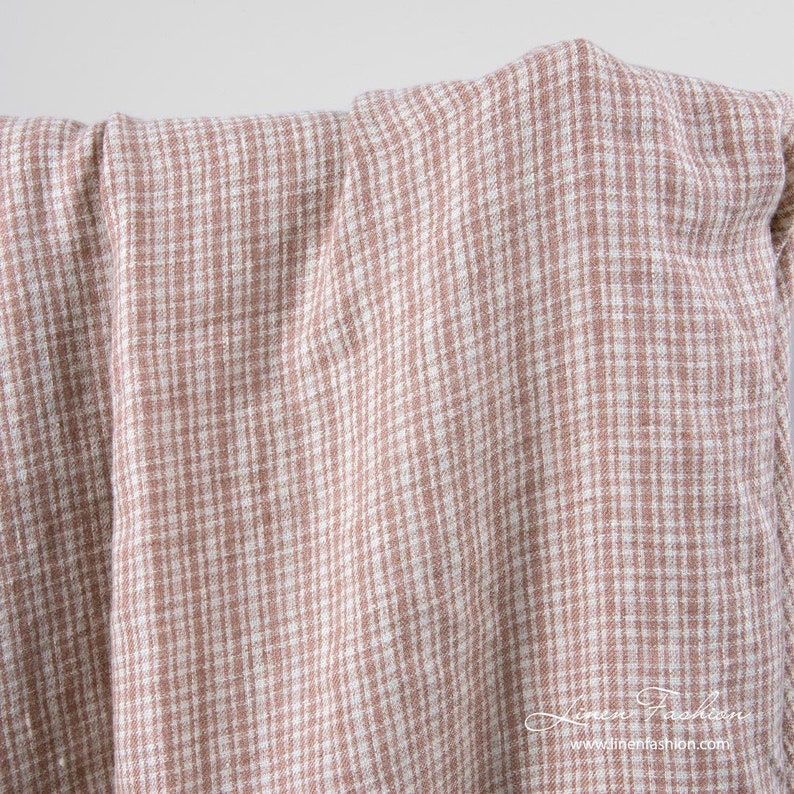 May include: A close-up shot of a folded fabric featuring a small, checkered design in light brown and white. The material appears to be linen or a similar natural fabric, with a slightly textured surface. The overall aesthetic is soft and casual.