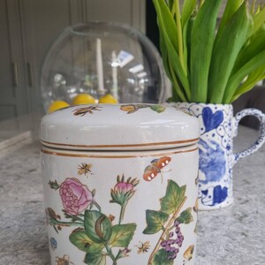 May include: A white ceramic jar with a lid, decorated with a floral design featuring pink roses, green leaves, purple grapes, and butterflies. The jar has a gold trim around the lid and base.