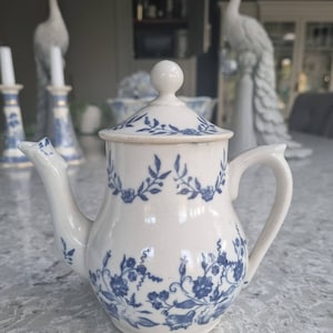 May include: A small white teapot with a blue and white floral pattern. The teapot has a lid and a handle.