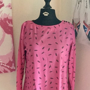 Pode incluir: Vestido rosa de manga comprida com padrão de penas pretas, exposto num manequim. O vestido tem decote redondo e comprimento pelo joelho. O manequim tem uma coroa. Uma almofada preta com texto na base.