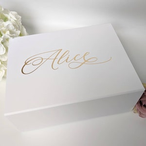 May include: A white gift box with the name "Alice" in gold script on the lid. The box is rectangular and closed. White flowers and a pink rose are in the background.