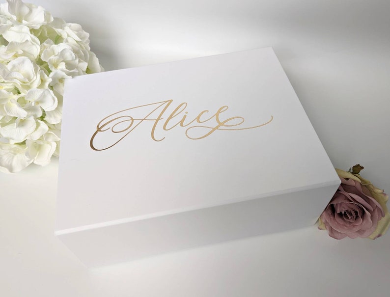 May include: A white gift box with the name "Alice" in gold script on the lid. The box is rectangular and closed. White flowers and a pink rose are in the background.