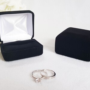 May include: A black velvet ring box, open and closed, with a white satin interior. Two silver rings with clear stones are in front of the boxes. The open box has a slot for the rings.