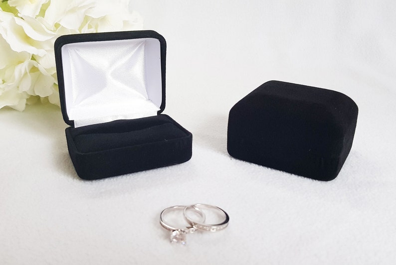 May include: A black velvet ring box, open and closed, with a white satin interior. Two silver rings with clear stones are in front of the boxes. The open box has a slot for the rings.