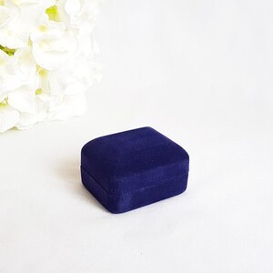 May include: A small, dark blue velvet ring box. The box is closed and sitting on a white surface.