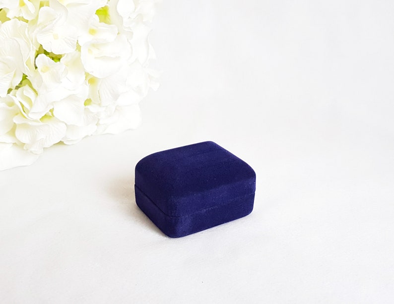 May include: A small, dark blue velvet ring box. The box is closed and sitting on a white surface.