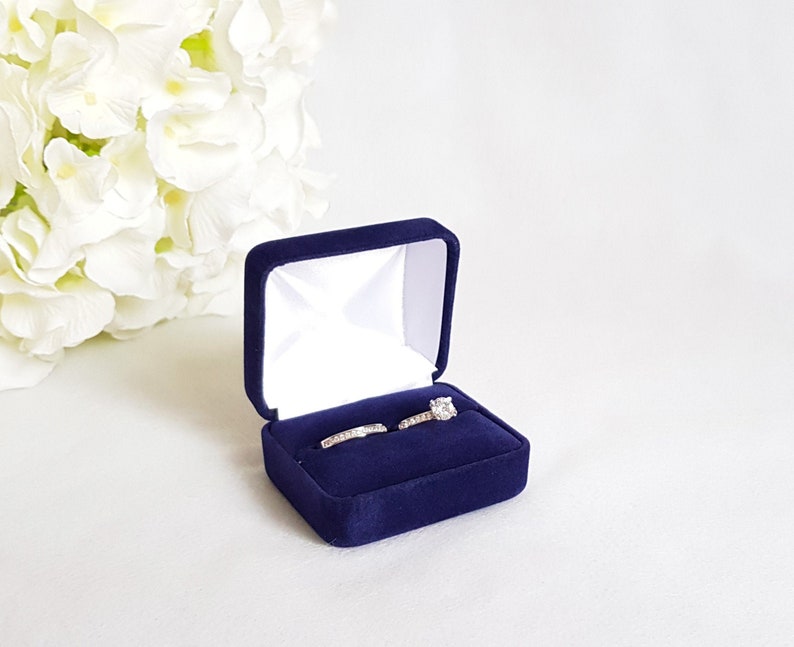 May include: A navy blue velvet ring box with two rings inside. The box is open and the rings are visible. The ring on the left is a simple band, and the ring on the right has a single stone.