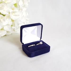 May include: A navy blue velvet ring box with a white interior. Two rings are displayed inside the box, one with a large centre stone and one with smaller stones.