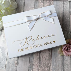 K&ouml;nnte beinhalten: Wei&szlig;e Geschenkbox mit goldener Schleife und Schleife. Die Box ist mit dem Text "Raheena The Beautiful Bride" in goldenen Buchstaben mit einem kleinen Herzen verziert.