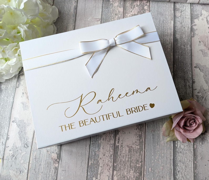 K&ouml;nnte beinhalten: Wei&szlig;e Geschenkbox mit goldener Schleife und Schleife. Die Box ist mit dem Text "Raheena The Beautiful Bride" in goldenen Buchstaben mit einem kleinen Herzen verziert.