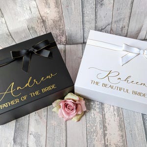 K&ouml;nnte beinhalten: Zwei Geschenkboxen mit personalisierten Botschaften. Die schwarze Box tr&auml;gt die Aufschrift "Andrew Father of the Bride" in goldenen Buchstaben. Die wei&szlig;e Box tr&auml;gt die Aufschrift "Raheena The Beautiful Bride" in goldenen Buchstaben.