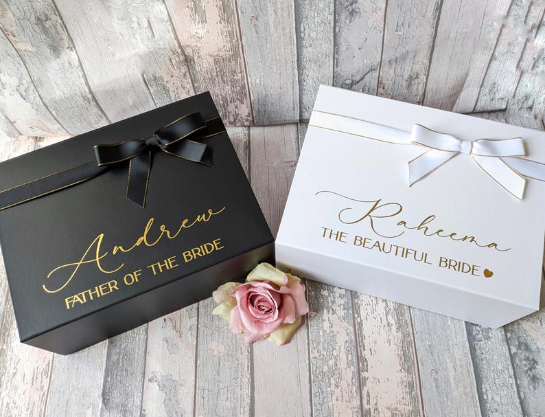 K&ouml;nnte beinhalten: Zwei Geschenkboxen mit personalisierten Botschaften. Die schwarze Box tr&auml;gt die Aufschrift "Andrew Father of the Bride" in goldenen Buchstaben. Die wei&szlig;e Box tr&auml;gt die Aufschrift "Raheena The Beautiful Bride" in goldenen Buchstaben.