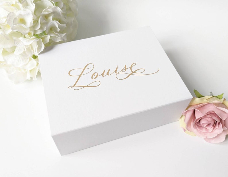 May include: A white gift box with the name "Louise" in gold script. The box is square and has a matte finish. White flowers and a pink rose are in the background, creating a soft, elegant aesthetic.