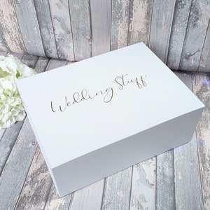 May include: A white, rectangular box with the words "Wedding Stuff" in gold script on the lid. The box is closed and sits on a wooden surface with a floral arrangement in the background.