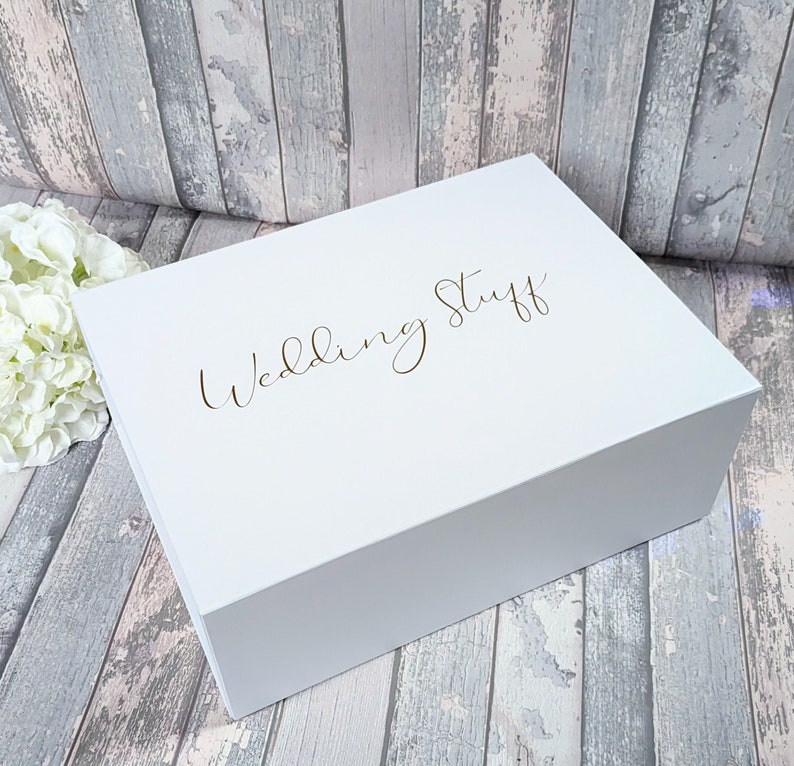 May include: A white, rectangular box with the words "Wedding Stuff" in gold script on the lid. The box is closed and sits on a wooden surface with a floral arrangement in the background.