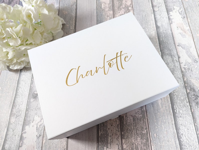 May include: A white gift box with the name "Charlotte" in gold script. The box is closed and sits on a wooden surface. A bouquet of white flowers is in the background.