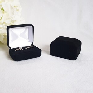 May include: Black velvet ring box, open to reveal two rings with clear stones. The box is lined with white satin. A second, closed black velvet box sits to the right. White flowers are in the background.