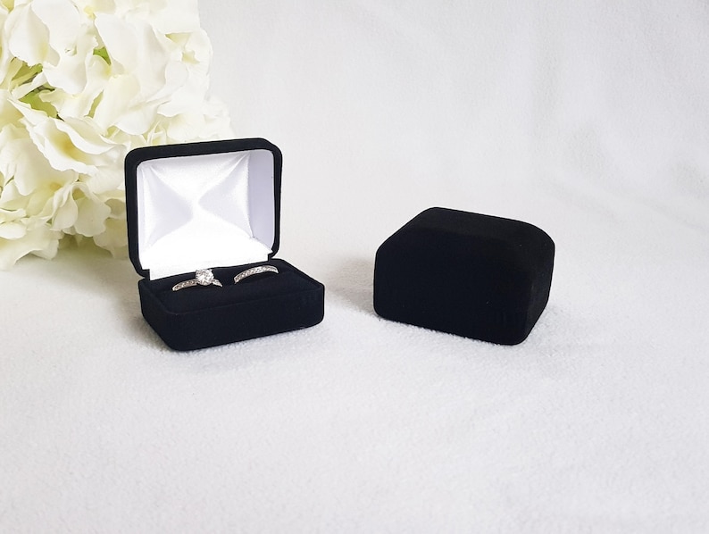 May include: Black velvet ring box, open to reveal two rings with clear stones. The box is lined with white satin. A second, closed black velvet box sits to the right. White flowers are in the background.