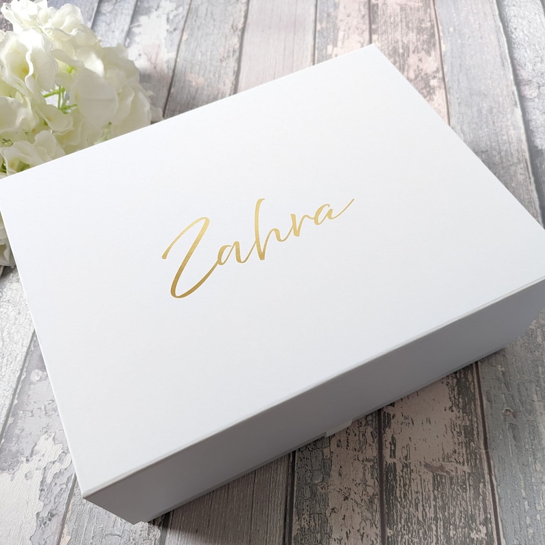 May include: A white gift box with a gold script font that reads "Zahra" on the lid. The box is rectangular and appears to be made of a matte material. White flowers are in the background.