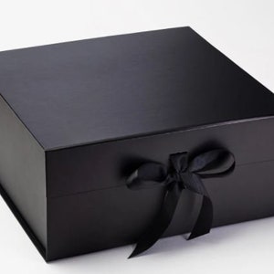 Puede incluir: Una caja de regalo negra con una cinta de satén atada en un lazo. La caja está hecha de cartón resistente y tiene un acabado liso y mate.