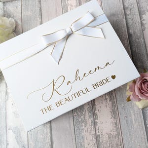 Può includere: Scatola regalo bianca con un nastro e un fiocco dorati. La scatola è decorata con il nome "Raheema" e il testo "The Beautiful Bride" in lettere dorate. Un piccolo cuore dorato è accanto al testo.