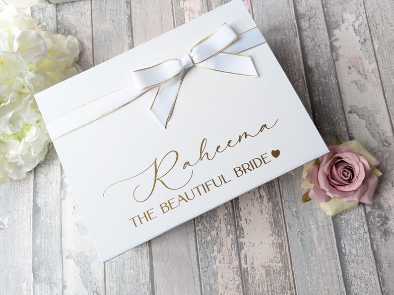 K&ouml;nnte beinhalten: Wei&szlig;e Geschenkbox mit goldener Schleife und Schleife. Die Box ist mit dem Namen "Raheema" und dem Text "The Beautiful Bride" in goldenen Buchstaben verziert. Ein kleines goldenes Herz befindet sich neben dem Text.