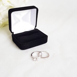 May include: A black velvet ring box with a white satin interior is open, displaying two silver rings. One ring features a large, clear gemstone, while the other is adorned with smaller stones. White flowers are in the background.