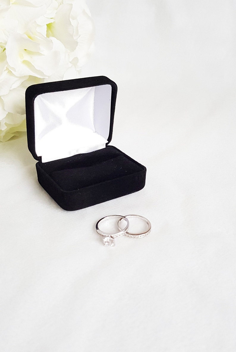 May include: A black velvet ring box with a white satin interior is open, displaying two silver rings. One ring features a large, clear gemstone, while the other is adorned with smaller stones. White flowers are in the background.