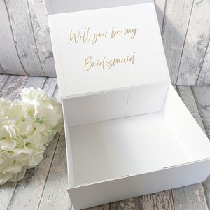 May include: A white gift box with a magnetic closure, open to reveal the inside. The lid features gold text that reads, "Will you be my Bridesmaid." A white floral arrangement sits to the left of the box.