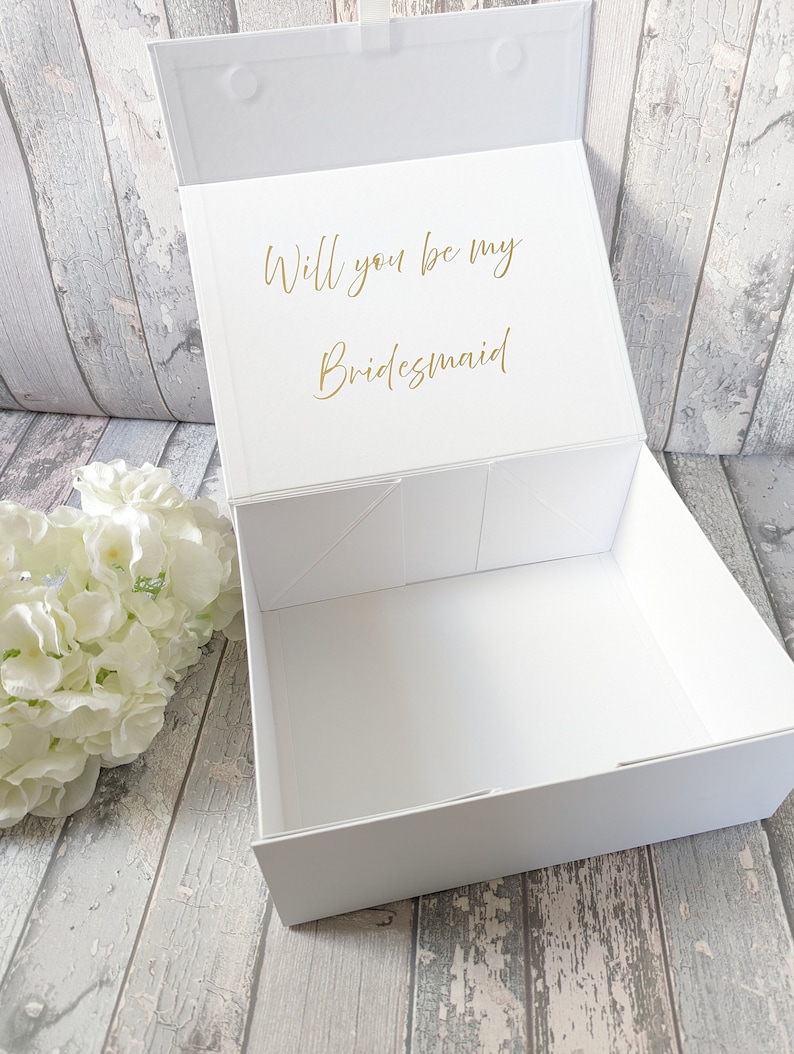 May include: A white gift box with a magnetic closure, open to reveal the inside. The lid features gold text that reads, "Will you be my Bridesmaid." A white floral arrangement sits to the left of the box.