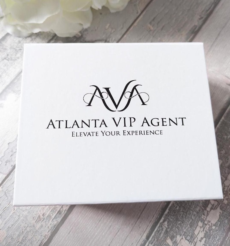 May include: A white box with the text "AVA ATLANTA VIP AGENT ELEVATE YOUR EXPERIENCE" in black. The box has a logo with the letters "AVA" in a stylized font. The background is a light gray wood-grain pattern.