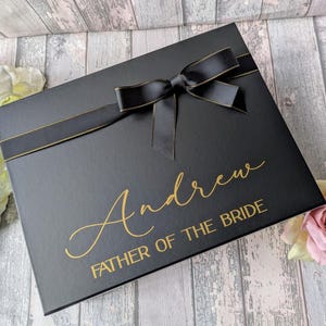 K&ouml;nnte beinhalten: Schwarze Geschenkbox mit einem schwarzen Satinband und Schleife. Die Box ist mit goldenen Lettern personalisiert, die "Andrew Father of the Bride" lauten.