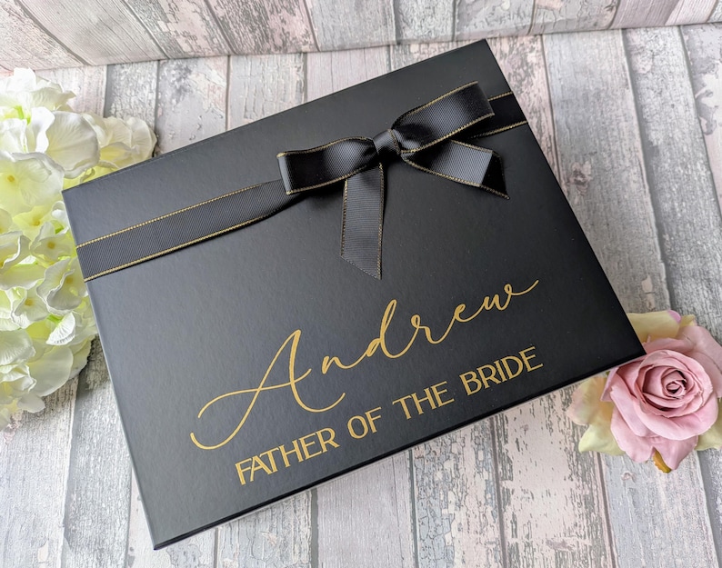 K&ouml;nnte beinhalten: Schwarze Geschenkbox mit einem schwarzen Satinband und Schleife. Die Box ist mit goldenen Lettern personalisiert, die "Andrew Father of the Bride" lauten.