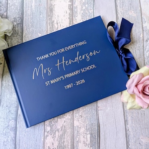 May include: A navy blue book with gold text reading "THANK YOU FOR EVERYTHING Mrs Henderson ST MARY'S PRIMARY SCHOOL 1997-2026." A navy ribbon and a pink rose are attached to the book.
