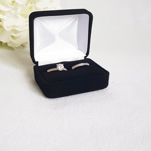 May include: A black velvet ring box with a white satin interior, holding two silver rings. One ring features a large, round, clear gemstone, and the other is a silver band with small, clear stones. The box is open, displaying the rings against a white background.