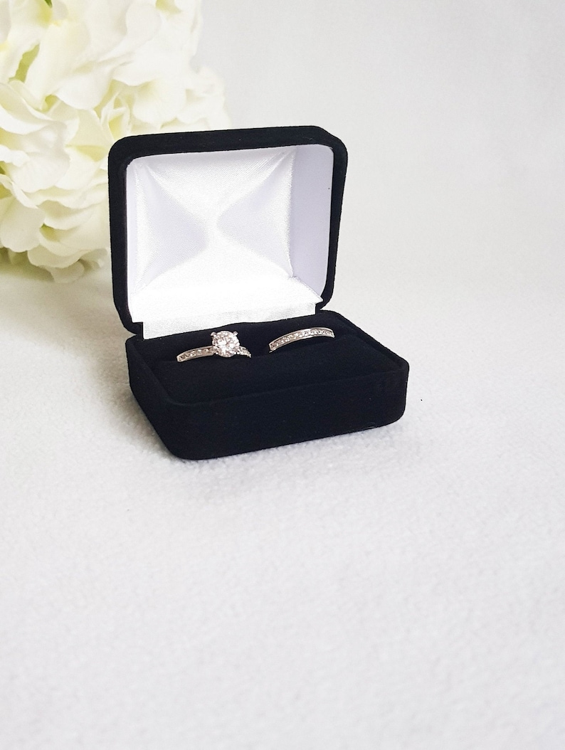 May include: A black velvet ring box with a white satin interior, holding two silver rings. One ring features a large, round, clear gemstone, and the other is a silver band with small, clear stones. The box is open, displaying the rings against a white background.