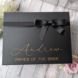 K&ouml;nnte beinhalten: Schwarze Geschenkbox mit goldener Schleife und Schleife. Die Box ist mit dem Namen "Andrew" und dem Text "Father of the Bride" in goldenen Buchstaben personalisiert.