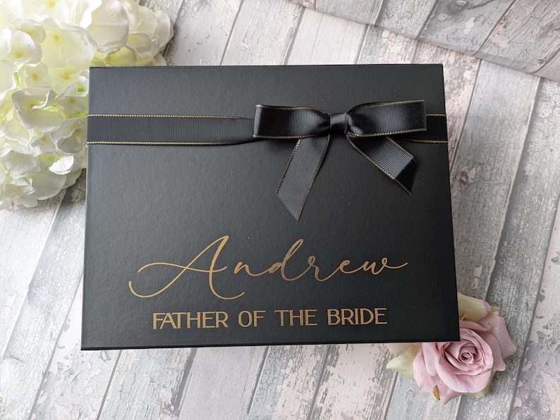 K&ouml;nnte beinhalten: Schwarze Geschenkbox mit goldener Schleife und Schleife. Die Box ist mit dem Namen "Andrew" und dem Text "Father of the Bride" in goldenen Buchstaben personalisiert.