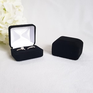 May include: Black velvet ring box, open to reveal two rings with clear stones. The box is square with a white satin interior. A second, closed black velvet box is next to the open box. White flowers are in the background.