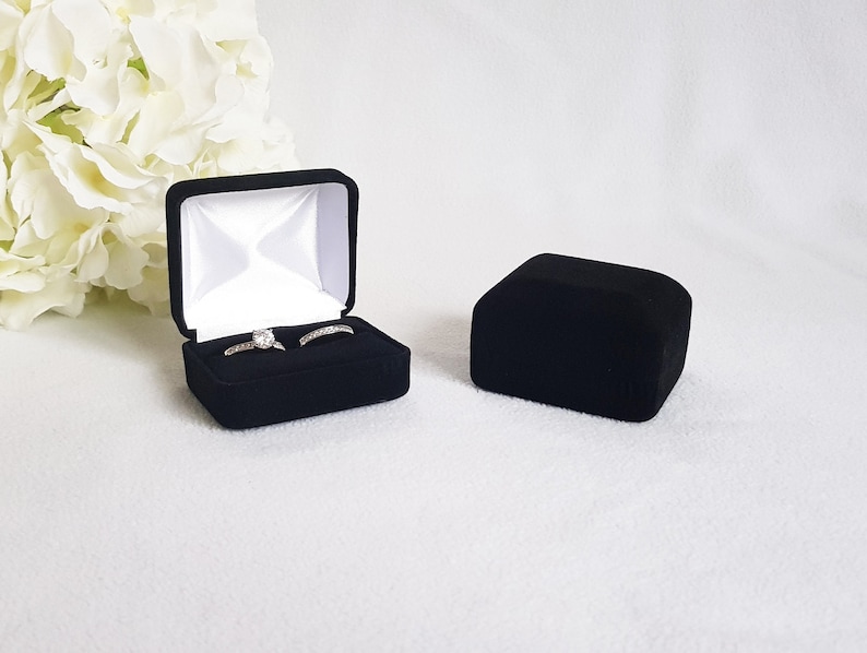 May include: Black velvet ring box, open to reveal two rings with clear stones. The box is square with a white satin interior. A second, closed black velvet box is next to the open box. White flowers are in the background.