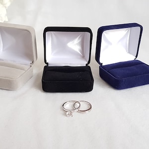 May include: Three ring boxes, one grey, one black, and one navy blue, are lined up on a white surface. Two silver rings are in front of the boxes.