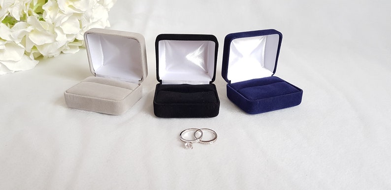 May include: Three ring boxes, one grey, one black, and one navy blue, are lined up on a white surface. Two silver rings are in front of the boxes.