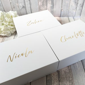 May include: Three white gift boxes with gold lettering. The names "Nicola", "Zahra", and "Charlotte" are written on the boxes. The boxes are on a wooden surface with a white flower arrangement in the background.
