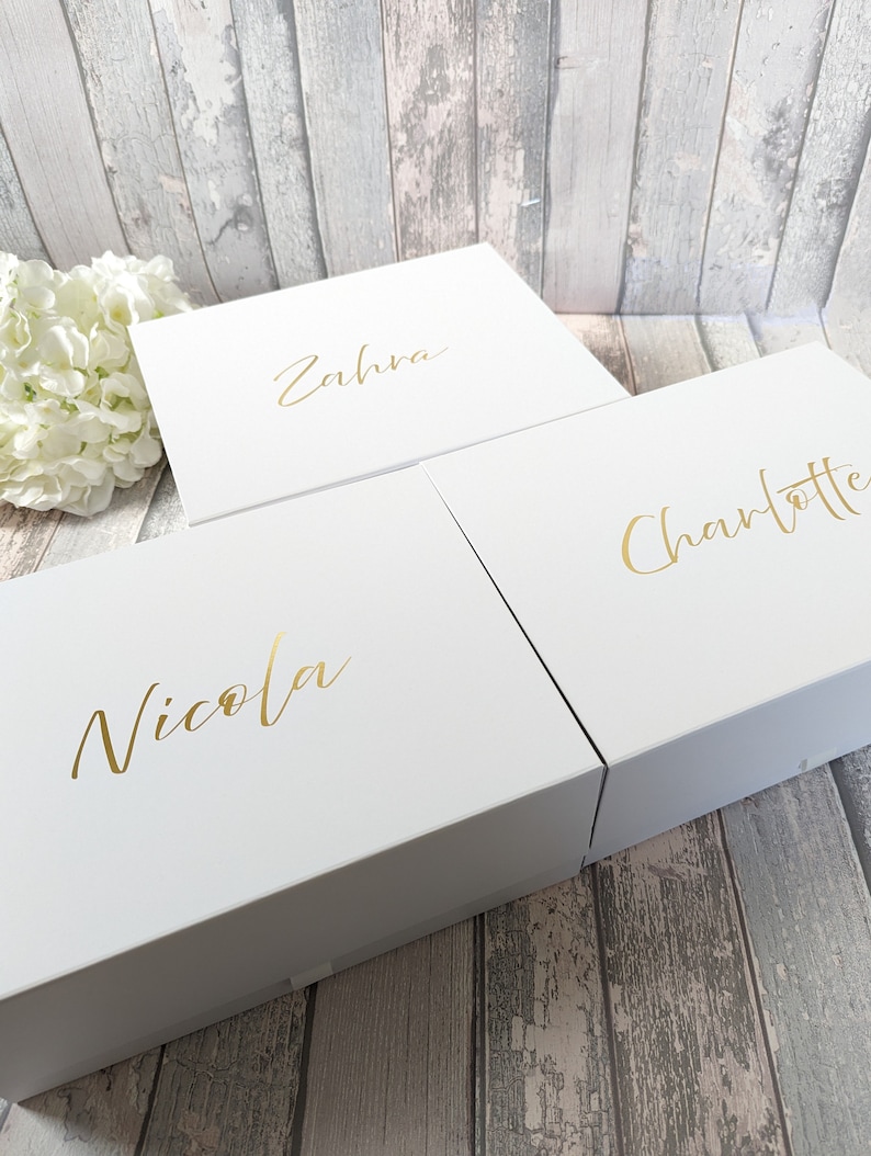 May include: Three white gift boxes with gold lettering. The names "Nicola", "Zahra", and "Charlotte" are written on the boxes. The boxes are on a wooden surface with a white flower arrangement in the background.