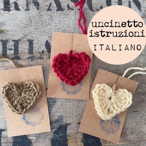 May include: Three handmade crochet heart ornaments in natural, red, and cream colors, attached to brown tags. The tags have a blue stamp and are displayed on a rustic wooden surface. Text in Italian is visible.
