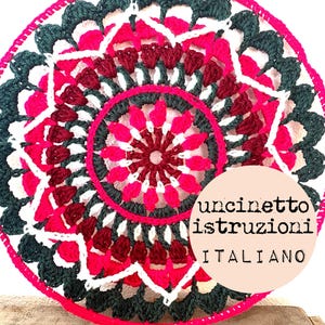 May include: A crocheted mandala with a pink, white, and dark green color scheme. The mandala is circular and has a repeating pattern of flowers. The text "uncinetto istruzioni ITALIANO" is written in white on a pink circle.